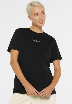 Blonde Person trägt ein schwarzes "Vivid Fusion" T-Shirt und beige Hose, schaut leicht nach rechts, die Hände locker vor dem Körper verschränkt.