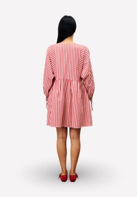 Frau mit langen schwarzen Haaren, die ein rot-weiß gestreiftes Kleid mit vertikalen Streifen und rote Schuhe trägt, steht mit dem Rücken zur Zuschauerin.