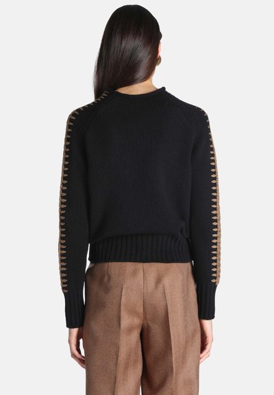 Femme aux longs cheveux foncés portant un pull noir avec des détails cousus beiges sur les manches et un pantalon marron taille haute, vue de dos.