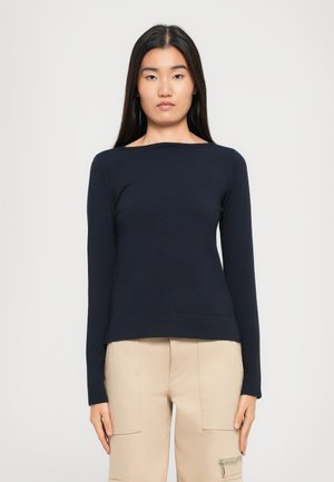 Jeune femme aux cheveux longs et raides portant un haut à manches longues bleu marine et un pantalon cargo beige, se tenant devant un fond clair uni.