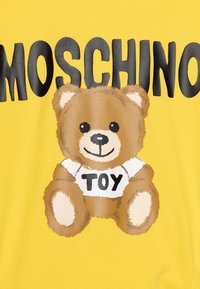 Il tessuto di cotone giallo presenta un grande orso dei cartoni animati con pelliccia marrone e una maglietta bianca con la scritta "TOY", insieme a bold lettere nere che formano la parola "MOSCHINO."