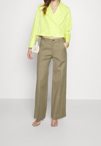 Cropped neon yellow jacket, double-breasted design, paired with high-waisted olive green wide-leg trousers, held with one hand. Clear heeled sandals.