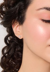 Kwadratowe kolczyki wkrętki z różowego złota z przejrzystym skupiskiem kamieni, solidnie osadzone, doskonale komponujące się z kręconymi włosami i gładką skórą.