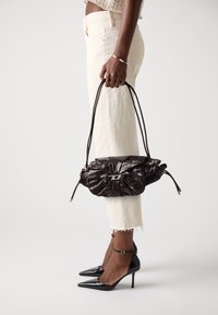 Dark brown leather handbag held by person wearing cream jeans, black pointed heels, and a beaded bracelet on wrist.