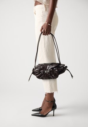 Sac en cuir marron foncé tenu par une personne portant un jean crème, des talons aiguilles noirs et un bracelet en perles au poignet.