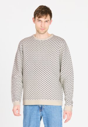 Lys beige sweater med mønster af sorte prikker, rund halsudskæring og ribbede manchetter. Båret med blå jeans, der viser en afslappet pasform.