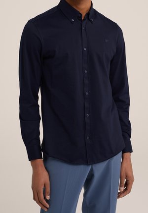 Mężczyzna ubrany w granatową koszulę na guziki z długim rękawem i kołnierzykiem oraz jasnoniebieskie spodnie, widoczny od szyi do bioder.