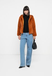 Oranje faux bont jas met een capuchon, zwarte top, lichtblauwe brede jeans en een kleine zwarte handtas. Vlakke schoenen maken de outfit compleet.