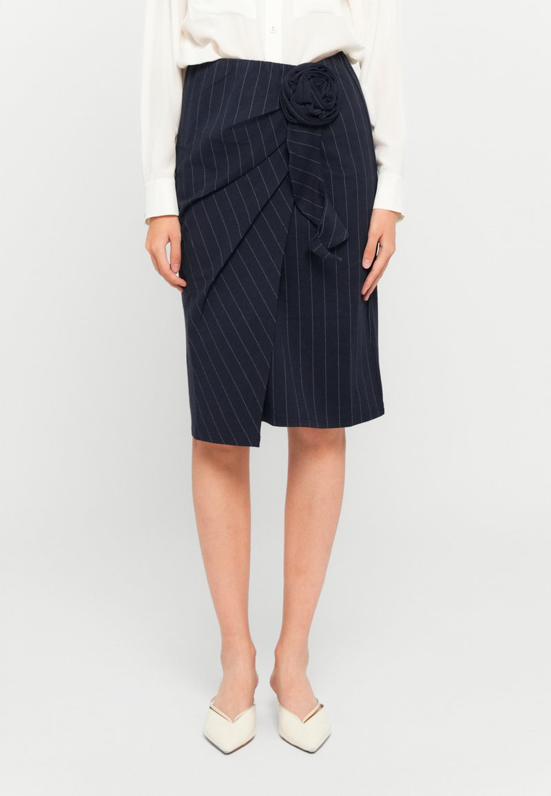 Vrouw in een marineblauwe pinstripe knielange rok met een voorste drapering en stoffen roosdetails, gecombineerd met een witte blouse en witte puntige schoenen.