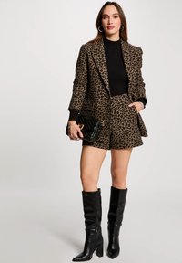 Ensemble composé d'un blazer et d'un short à imprimé léopard marron, associé à un pull côtelé noir et des bottes noires à talons hautes jusqu'aux genoux. Sac à main noir matelassé en accessoire.