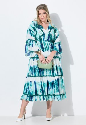 Maxikleid in Weiß mit blauen und grünen Aquarellstreifen, mit ausgestellten Ärmeln und rüschensaum. Kombiniert mit einer hellgrünen Handtasche und Absätzen.
