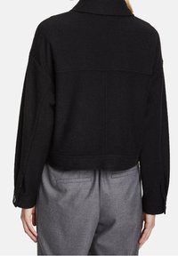 Žena oblečená v černé dlouhé bundě s manžetami na knoflíky a šedých kalhotách na míru, zobrazená ze zad na bílém pozadí.