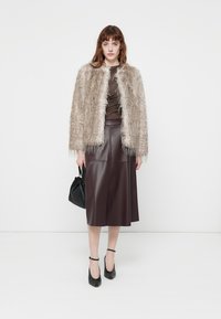 Woman wearing beige faux fur jacket, brown top, dark leather midi skirt, black ankle-strap heels, holding small black handbag, standing on white background.