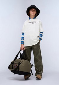 Ikdīgs apģērbs ar baltu grafisko T-kreklu virs zila svītrainā garām piedurknēm, olīvzaļām biksēm un melnu pogu cepuri, turot olīvzaļo duffel somu.