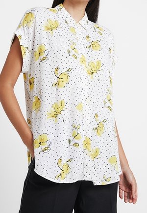 Chemise blanche à manches courtes boutonnée avec pois noirs et imprimé floral jaune, portée avec un pantalon noir.