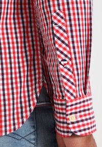 Camisa de cuadros rojos y blancos con detalles azules, puño abotonado y costuras contrastantes. La tela tiene una textura suave y un diseño entallado.