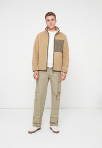 Junger Mann, der vor einem weißen Hintergrund steht. Er trägt eine beige Fleecejacke mit Brusttasche, ein weißes T-Shirt, Cargo-Hosen und weiße Sneakers.