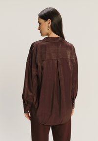 Mujer con cabello largo y oscuro que lleva una blusa suelta y brillante de color marrón oscuro con un pliegue central en la parte posterior y pantalones oscuros a juego, de perfil hacia la izquierda.