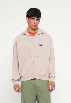 Light beige zip-up hoodie made of soft fabric, featuring a front pocket, drawstring hood, and a small multicolored logo on the chest.