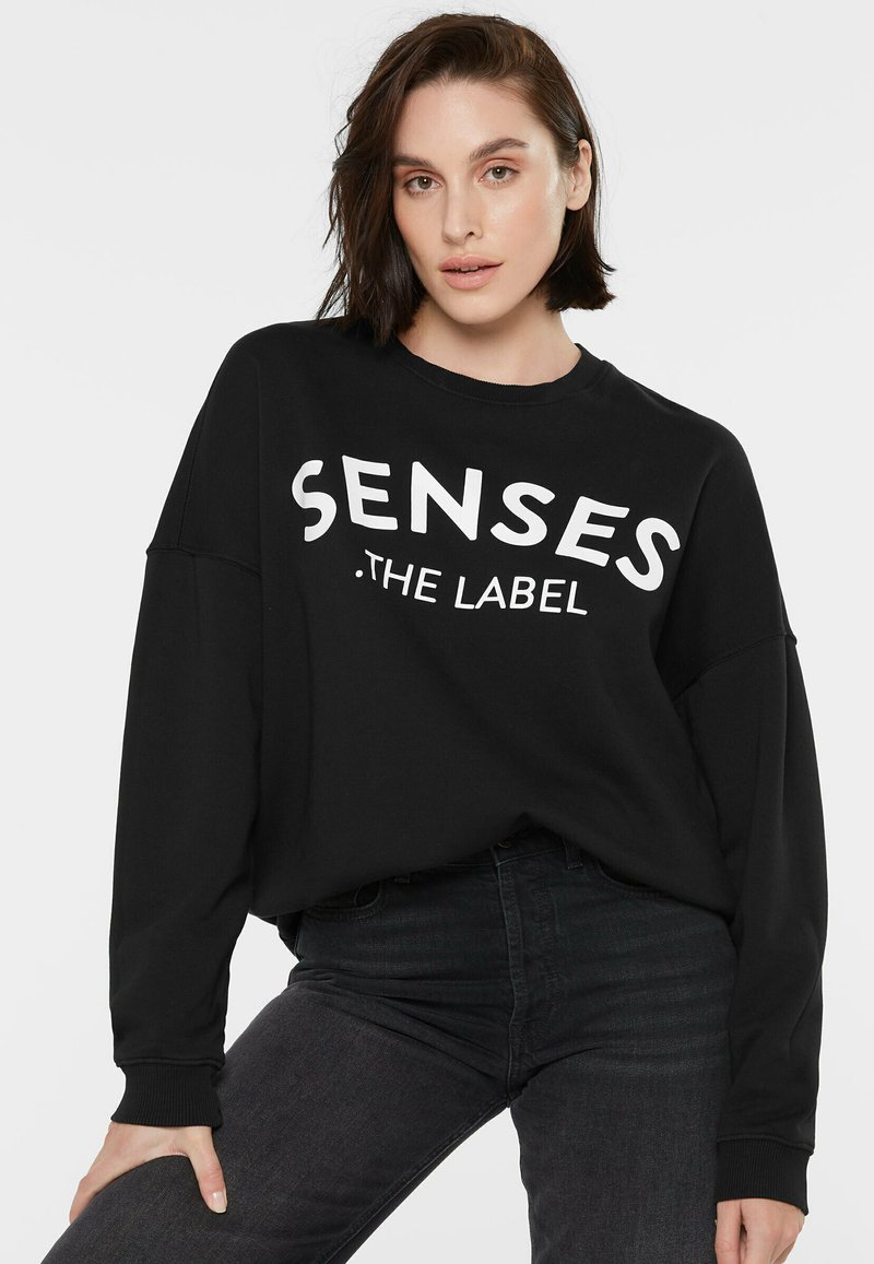 Schwarzes Sweatshirt mit langen Ärmeln, das auf der Vorderseite den weißen Text "SENSES .THE LABEL" trägt. Weicher Stoff mit lockerer Passform und Bündchen an den Handgelenken.