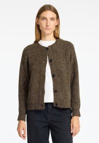 Brauner gestrickter Cardigan aus weichem Material, mit rundem Ausschnitt und sechs schwarzen Knöpfen auf der Vorderseite. Getragen über einem weißen Hemd.