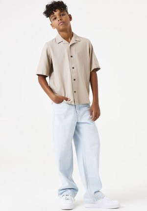 Camisa casual de manga corta en un beige claro, con tejido texturizado y delante abotonado. Combinada con pantalones de denim sueltos en azul claro y zapatillas blancas.