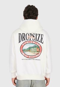 Person in einem weißen Hoodie mit einem bunten Strand- und Bergmotiv sowie dem Text "DROPSIZE Clothing Worldwide Luxury Streetwear" auf dem Rücken.