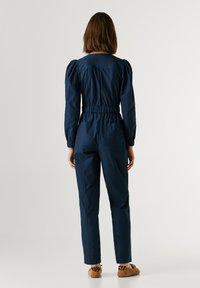 Ολόσωμη φόρμα σε ναυτικό μπλε με φουσκωτά μανίκια, ελαστική μέση και στενές μπατζάκια. Διαθέτει πίσω τσέπες και απαλό βαμβακερό ύφασμα.