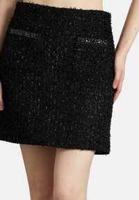 Falda corta de tweed negro con tejido texturizado, que presenta dos bolsillos delanteros adornados con acentos de cuentas. El dobladillo es recto y está acabado.