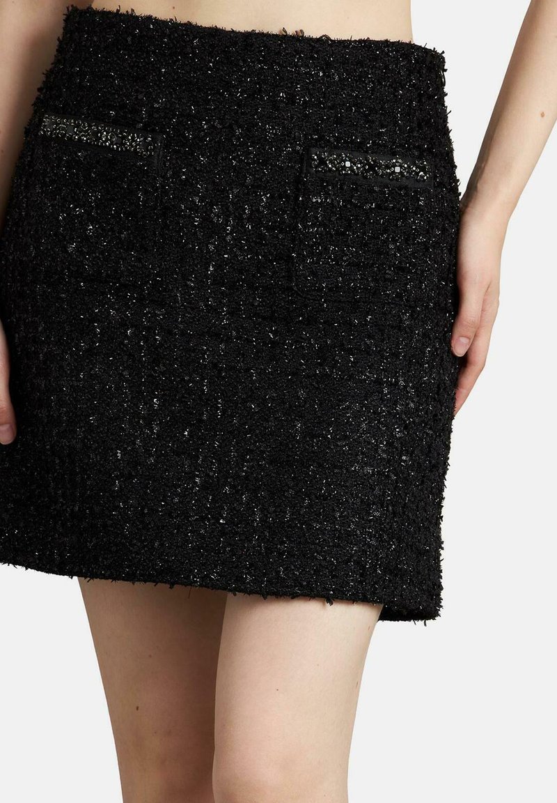Falda corta de tweed negro con tejido texturizado, que presenta dos bolsillos delanteros adornados con acentos de cuentas. El dobladillo es recto y está acabado.