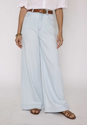 Donna che indossa jeans azzurri a gamba larga, cintura di pelle marrone, top bianco con maniche arricciate, braccialetti e sandali marroni.