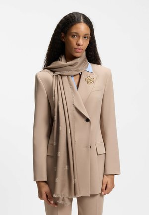 Mujer con cabello largo y rizado que lleva un traje beige, una bufanda a juego alrededor del cuello y un broche de flor dorada en la solapa.