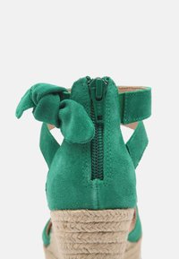 Sandale compensée en suède vert avec fermeture éclair au talon et nœud décoratif. Texture en jute tressé sur le talon compensé. Design avec talon et bride de cheville ouverts.