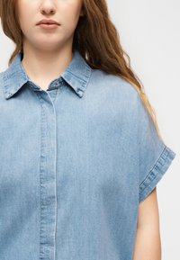Γυναίκα που φοράει ένα ανοιχτό μπλε πουκάμισο από τζιν με κοντά μανίκια και γιακά, στέκεται μπροστά σε ένα απλό ανοιχτό φόντο.