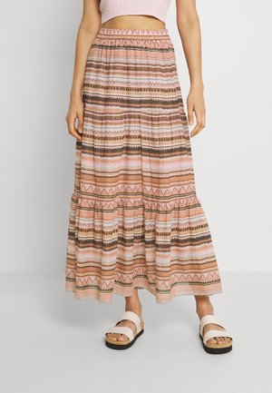 Kvinde iført en lyserød crop top, ankel-lang lagdelt nederdel med vandrette geometriske mønstre og hvide platform-sandaler, stående mod en ensfarvet baggrund.