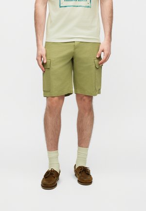 Homme portant un short cargo vert olive, des chaussettes crème et des chaussures bateau marron, debout devant un fond uni.