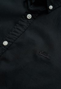 Camisa negra de botones con una textura suave, que presenta botones blancos y un logo bordado en negro en el lado izquierdo.