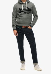 Sudadera gris con cordones y logo negro, camisa de cuadros debajo, vaqueros oscuros y zapatillas claras con puños remangados.