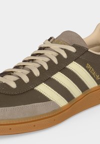 Καφέ αθλητικό παπούτσι Adidas Spezial με κρεμ τρεις ρίγες, μπεζ κορδόνια, υφασμένες πλευρές και σόλα από καουτσούκ σε ουδέτερο φόντο.