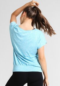Camisa de manga corta azul claro con un corte holgado, escote redondo y detalle fruncido en la parte posterior, combinada con leggings negros.