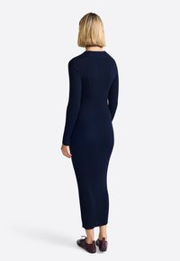 Femme aux cheveux blonds courts portant une robe midi côtelée bleu marine à manches longues et des chaussures en cuir bourgogne, se tenant de dos.