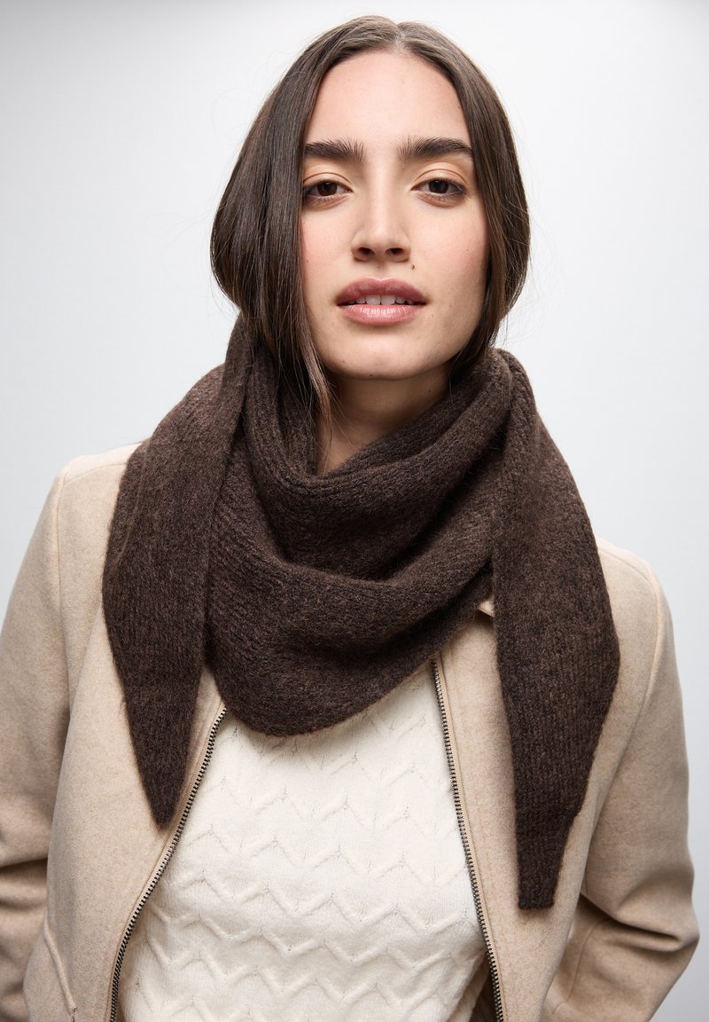Sciarpa di lana marrone a forma triangolare, con una texture morbida e bordi irregolari, appoggiata intorno al collo, abbinata a una giacca beige chiaro.