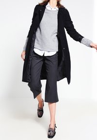 Kobieta idąca w czarnym długim płaszczu na szary sweter, białą koszulę, przycięte ciemne spodnie i czarne wzorzyste buty na obcasie.