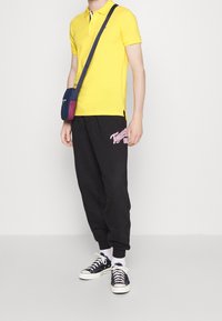 Polo shirt jaune, jogging noir avec logo rouge, et baskets noires. Design décontracté avec une coupe confortable et des couleurs contrastées.