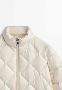 Veste matelassée beige crème avec col montant et fermeture éclair frontale, présentée sur un fond blanc uni.