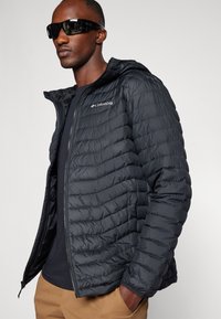Sort puffeblazer med quiltet design, lynlås lukning og hætte. Har et Columbia-logo på brystet, båret over en mørk skjorte.