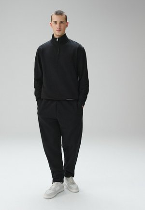 Young man with short hair wearing a black long-sleeve zip-up top, black pants, and white sneakers, standing with hands in pockets.