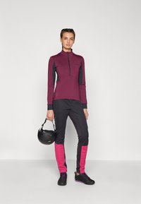 Athletisk zip-up topp i burgunder med sorte detaljer; sorte bukser med rosa paneler; modellen holder en svart hjelm. Skoene har lilla detaljer.