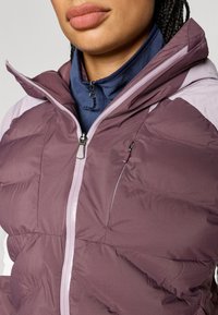 Vijolična puhasta jakna z visokim ovratnikom, z zadrgo spredaj in svetlimi poudarki. Ima stranski žep in teksturirano tkanino.