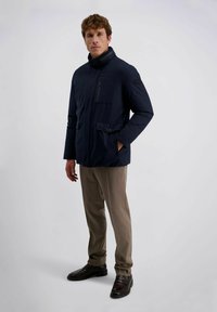 Donkerblauwe geïsoleerde jas met een hoge kraag, een ritssluiting aan de voorkant en meerdere zakken, gecombineerd met lichtbruine broek en donkere schoenen.